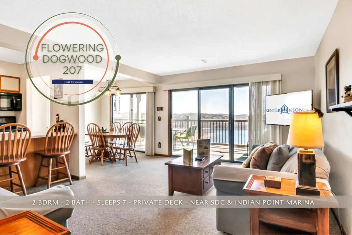 The image showcases a bright, inviting open-concept living space with a comfortable seating area, a spacious dining table perfect for group gatherings, and a kitchen bar for casual meals. Glass doors reveal a private deck with panoramic views overlooking Table Rock Lake, blending the warmth of the interior with the beauty of the outdoors.