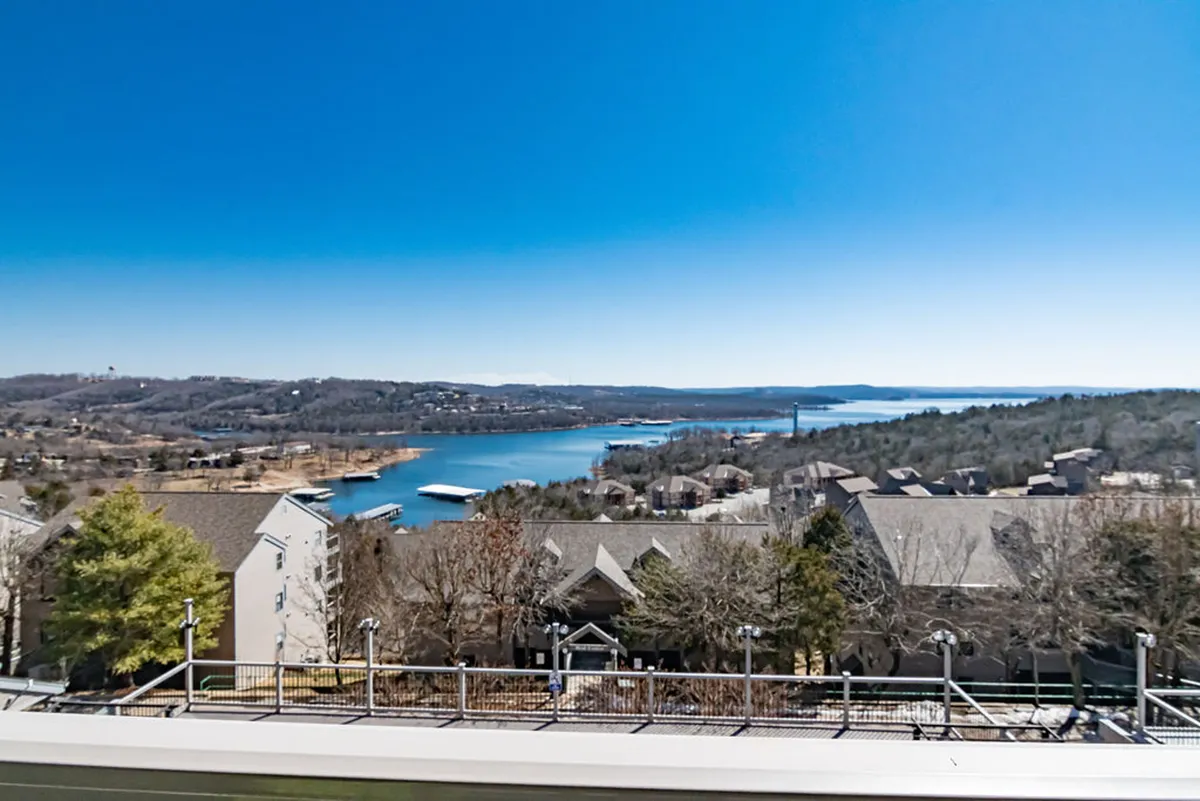 A tranquil view from a balcony or outdoor seating area, overlooking Table Rock Lake with rolling hills and neighborhood houses in the background. The scene captures the peaceful, natural beauty of the lakeside setting and suggests outdoor enjoyment.