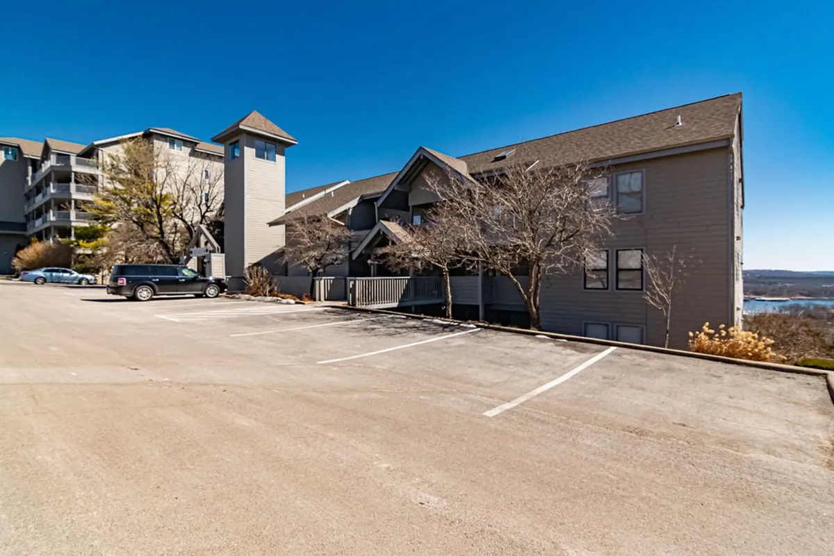 The image shows a multi-level building nestled near the lake, with a distinctive tower structure and a peaceful, natural setting. The bare trees and expansive parking area suggest a quiet retreat, and the lake nearby is visible, setting a tranquil scene perfect for unwinding while watching sunsets.