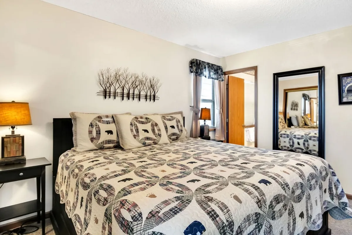 A spacious king bedroom featuring a bed with a patterned quilt, a nightstand with a lamp, decorative wall art, and a large mirror reflecting the room. The overall mood is warm and inviting, with soft lighting and thoughtful decor that create a restful sanctuary. The placement of the TV, though not directly visible, is implied by the comfortable setup for relaxed evenings.