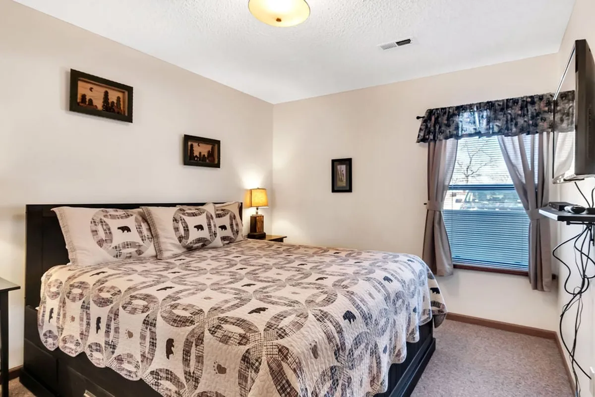 A cozy king bedroom with a plush patterned quilt, nightstand with a softly glowing lamp, framed artwork on the walls, and a window with curtains. The overall setting is serene and intimate, perfect for reading or unwinding before bed. The TV is implied by the room's setup for relaxed evenings.