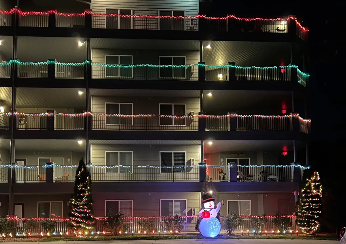 An outdoor scene featuring cheerful string lights, a playful snowman decoration, and a brightly lit Christmas tree. The area is set up for gatherings, celebrations, and group enjoyment, suggesting a lively and inviting outdoor environment.