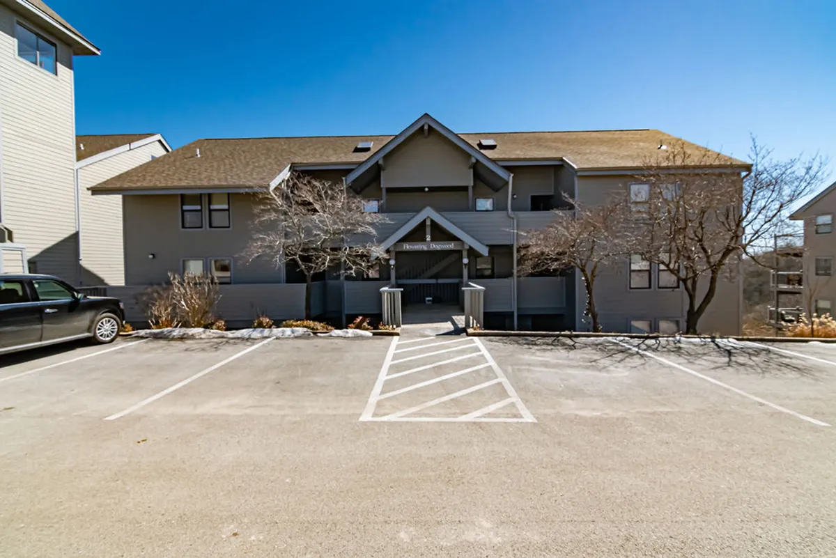 The welcoming exterior of the apartment building, complete with an entrance awning and framed by mature trees, offers a sense of arrival and accessibility. The entrance is easily visible from the parking area, hinting at a stair-free, convenient walk-in layout.
