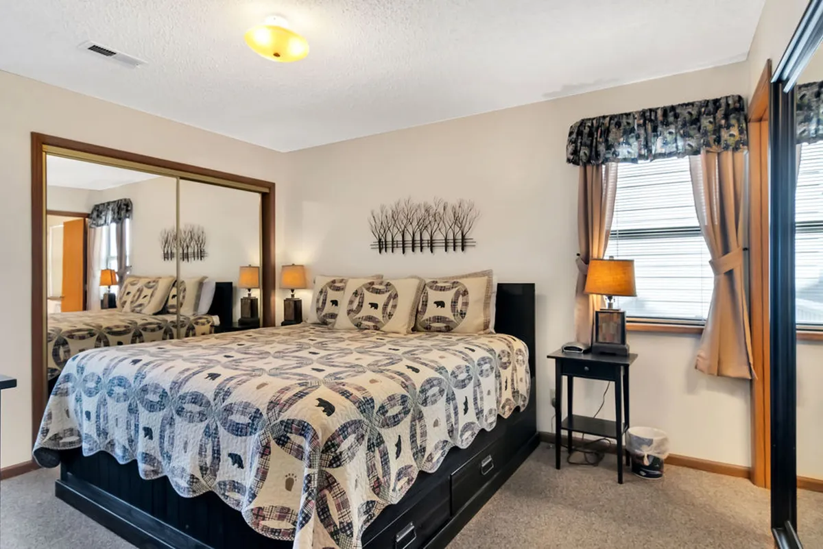 Another angle of a king bedroom, highlighting a bed with a patterned quilt, mirrored dresser, and wooden furniture. The natural light streaming in from the windows enhances the welcoming ambiance, while the mirror adds depth to the space. The room exudes comfort and tranquility, ideal for guests seeking a peaceful retreat.