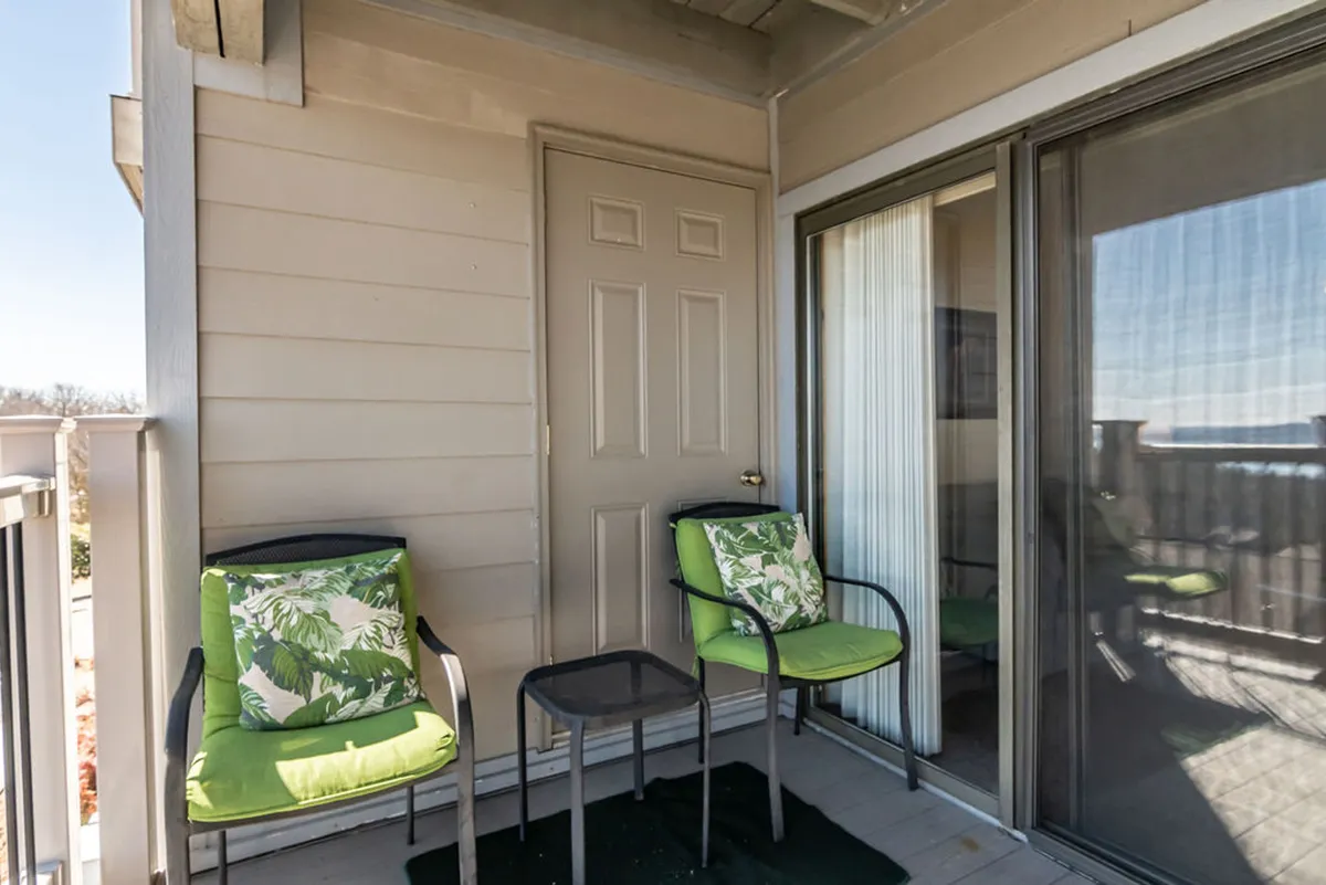 A tranquil private deck with green chairs featuring floral cushions, a small black table set between them, and a door leading back into the home. The setting hints at a relaxing morning or peaceful evening, with space to enjoy coffee while overlooking the natural surroundings.