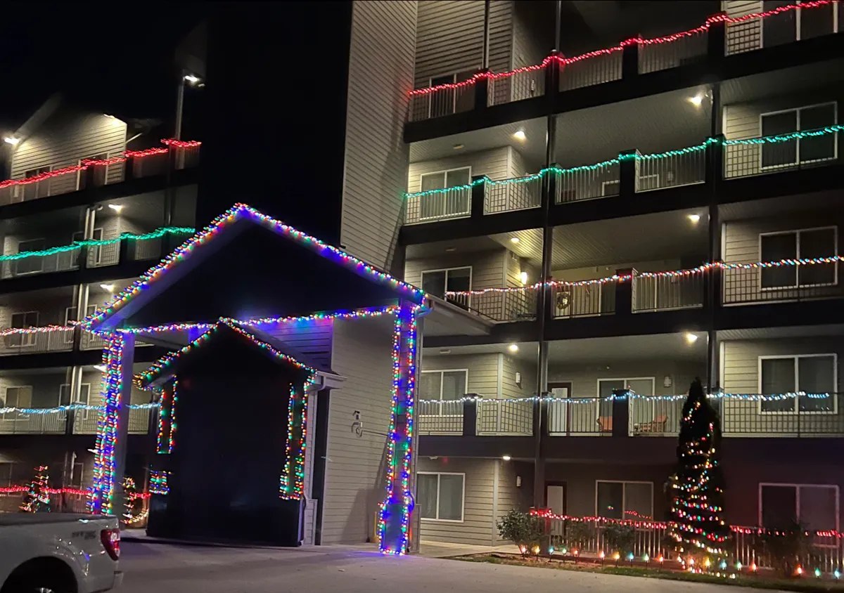 The image shows the exterior of the building adorned with vibrant Christmas lights, a warmly decorated entrance, and a glowing Christmas tree, all set against a festive evening backdrop. The scene captures a sense of arrival and community celebration, hinting at seasonal gatherings and the lively atmosphere awaiting guests.