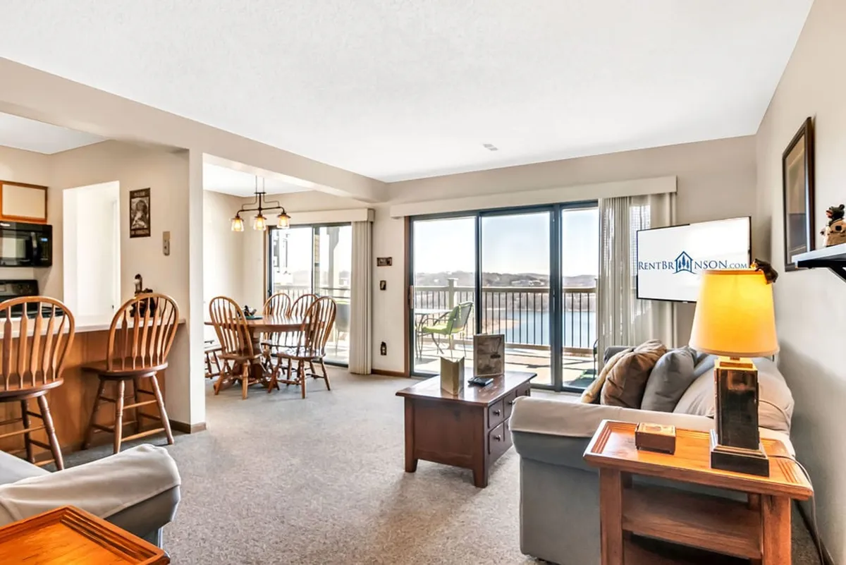 This inviting open-concept living and dining area features a large window framing stunning lake views. Comfortable seating is arranged for conversation or movie nights, with a dining table nearby for shared meals. The space is filled with natural light and seamlessly connects to the private deck beyond.