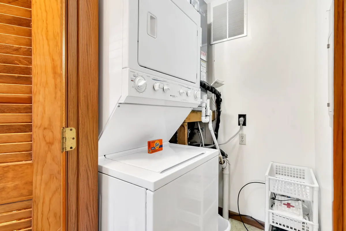This image showcases a stacked washer and dryer neatly set within a laundry area, alongside a wooden door and a laundry basket. The space feels organized and is clearly dedicated to laundry tasks, emphasizing convenience and efficiency for guests.