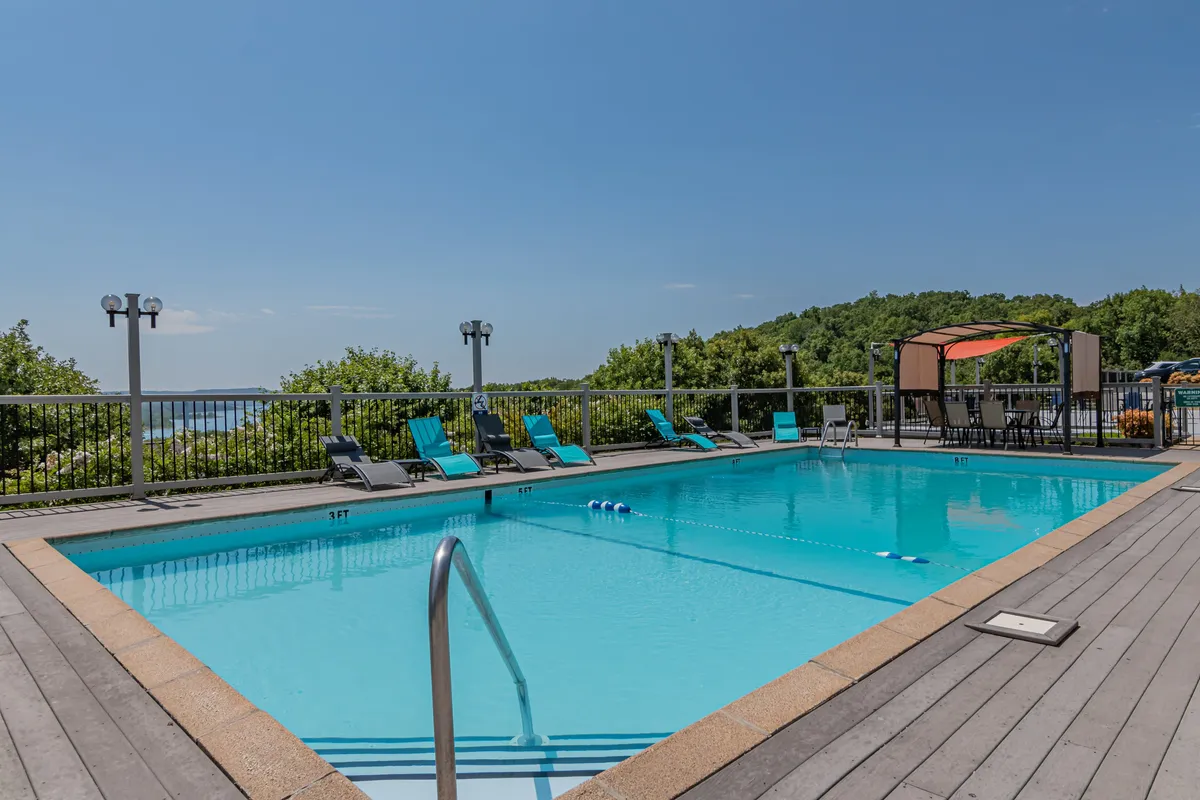 A sun-drenched resort pool surrounded by lounge chairs, a shaded canopy area, and lush landscape views. The pool area invites both relaxation and socializing in a clean, comfortable outdoor setting.